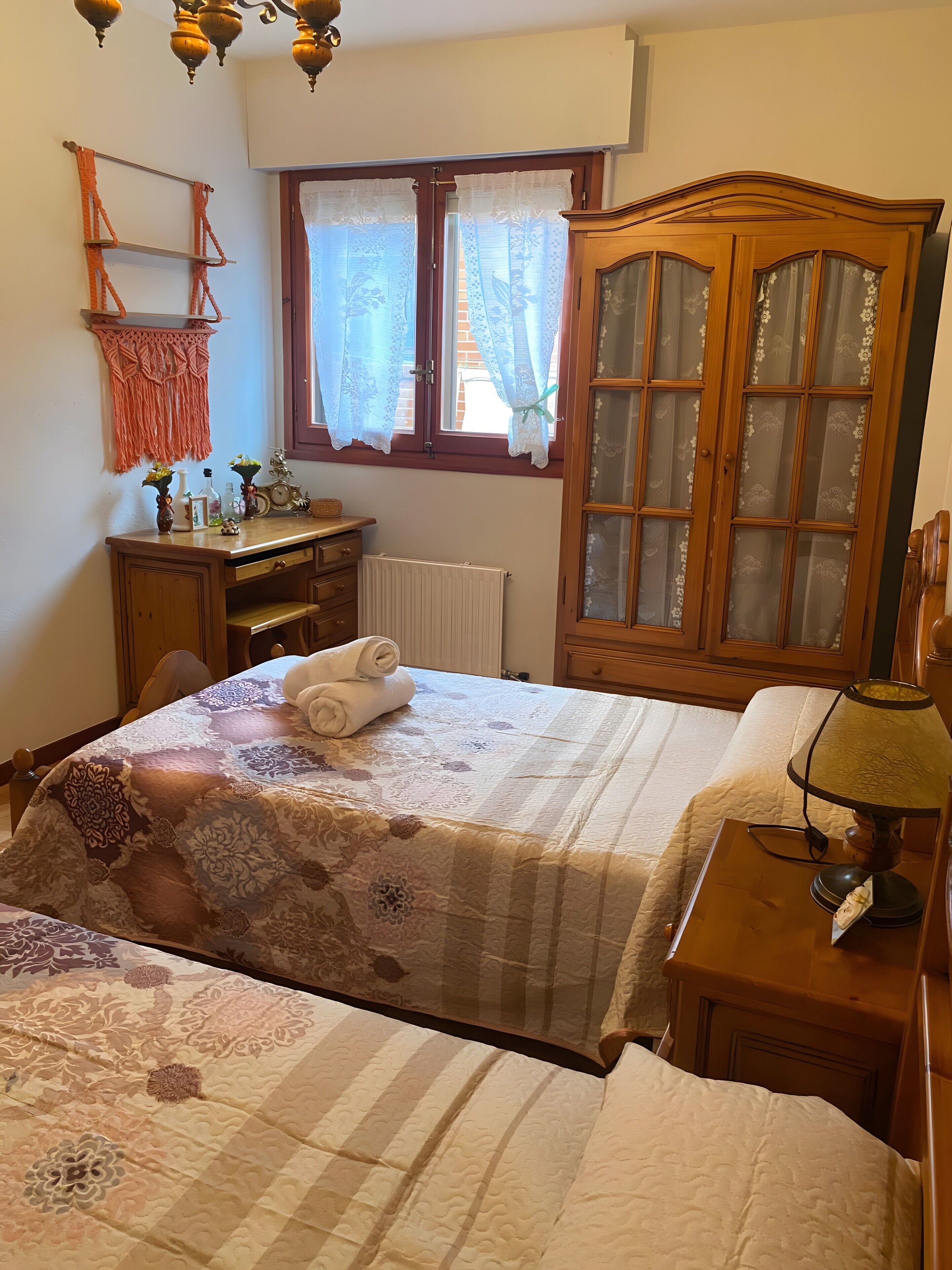 Un dormitorio con dos camas, un armario de madera, un escritorio con silla, un radiador y una ventana con cortinas.