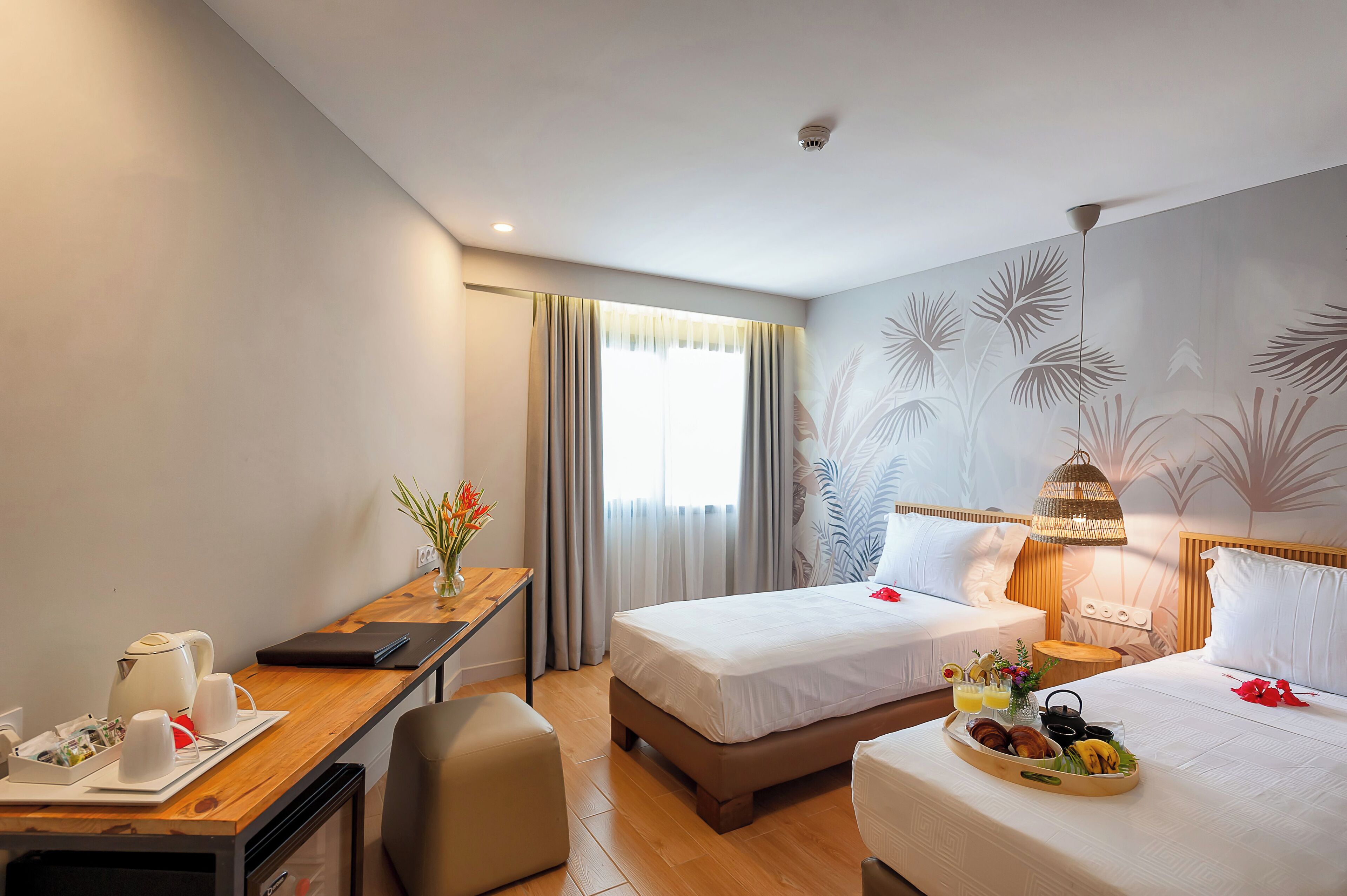 A hotel room with two beds, a desk with breakfast items, a small table with flowers, and a wall with a leaf pattern.