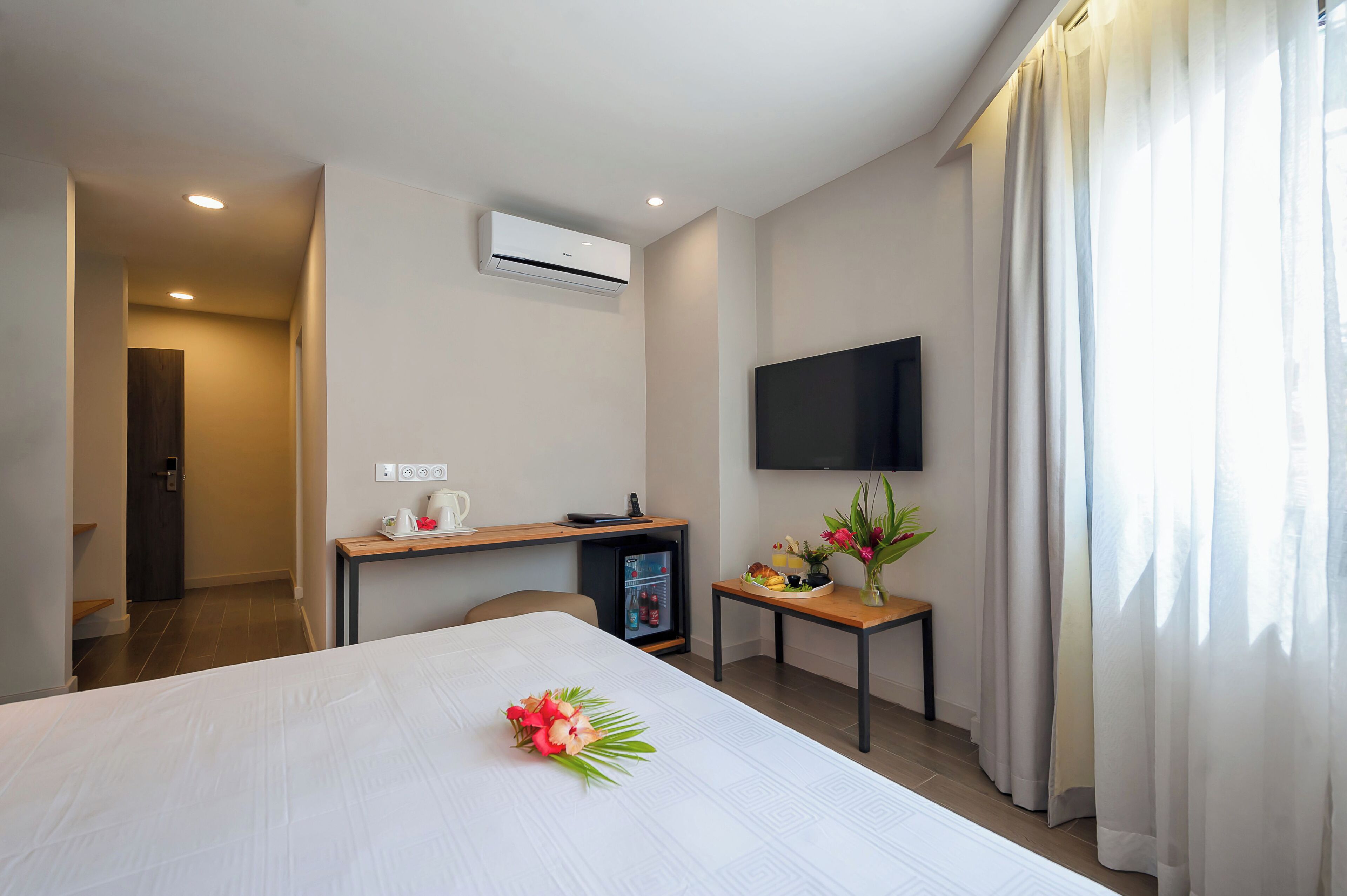 A hotel room with a bed, a desk with a computer, a flat-screen TV, an air conditioner, and a window with curtains.