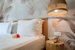 Una cama bien hecha con sábanas blancas y una sola flor roja, un lámpara de fibras tejidas en el techo, y una pequeña mesita de madera con libros.
