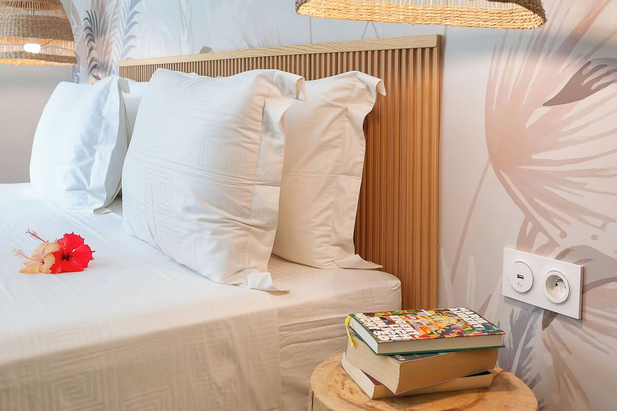 Ein Schlafzimmer mit Bett, weißer Bettwäsche, einem Holz-Kopfteil, einer gewobenen Pendelleuchte und einem kleinen Holzschemel mit einem Buch.
