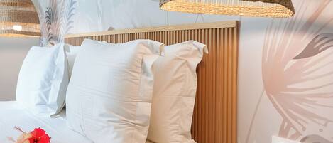 A bedroom with a bed, white bedding, a wooden headboard, a woven pendant light, and a small wooden stool with a book.