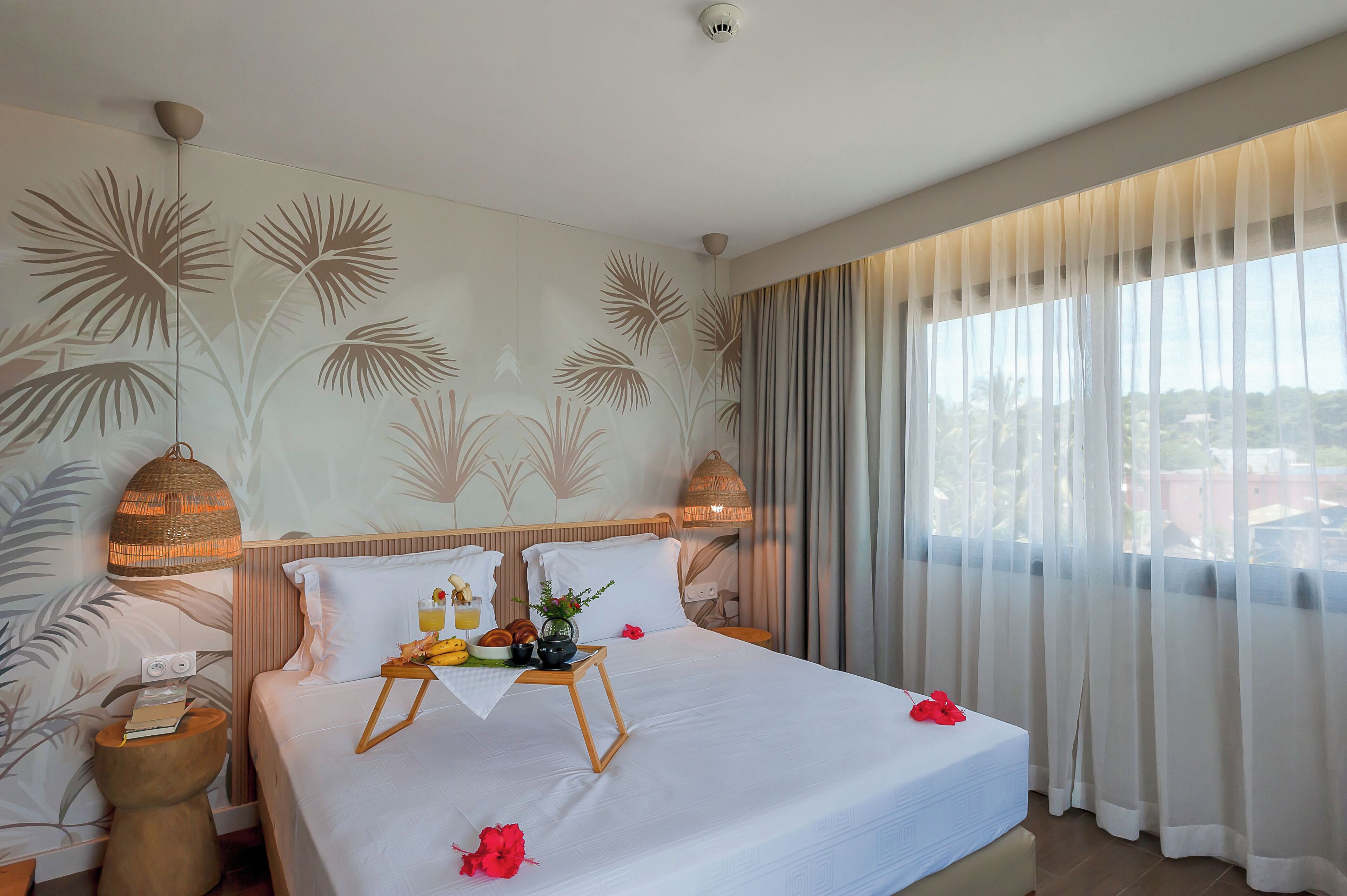 Une chambre d’hôtel avec un lit, une table de chevet avec une lampe, une fenêtre avec des rideaux légers et un mur orné d’un motif de feuilles de palmier.