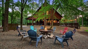 Eine Hütte mit Feuerstelle, umgeben von Adirondack-Stühlen, eingebettet in eine bewaldete Gegend mit einer Holzterrasse und Lichterketten.