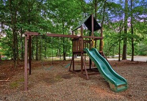 A playground with a slide and swings set in a wooded area. - Playset | Outdoor Games | Hot Tub | Fire Pit (Broken Bow)