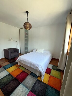 Ein Schlafzimmer mit einem Bett, einem grauen Sessel, einer weißen Lampe und einem farbenfrohen Teppich.