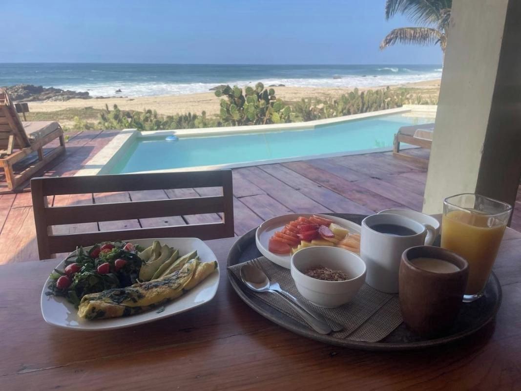 Bandeja de desayuno con comida y bebidas en una terraza de madera con vista a una playa y al océano.