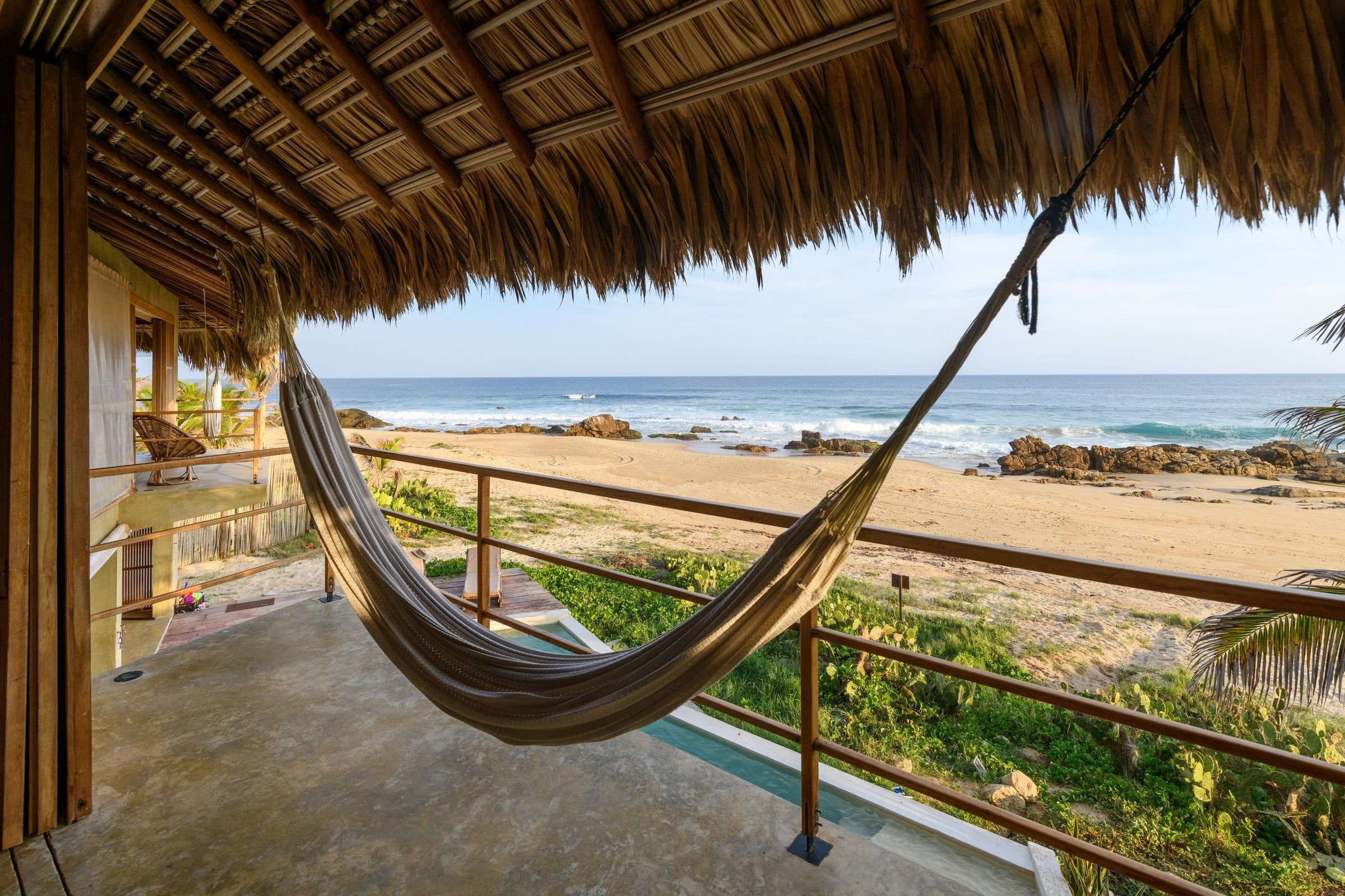 A hammock on a balcony overlooking a beach with a thatched roof structure.