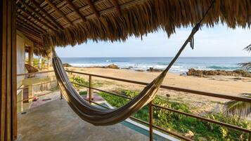 A hammock on a balcony overlooking a beach with a thatched roof structure.