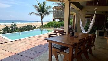 Une terrasse en bois avec une piscine, une table à manger et des chaises, donnant sur une plage bordée de palmiers et sur l’océan.