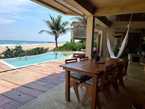 A wooden deck with a pool, a dining table, and chairs, overlooking a beach with palm trees and the ocean. - Las Rocas: beach front room with infinity pool and spectacular views (Agua Blanca, Tonameca)
