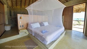 Ein Schlafzimmer mit einem Bett unter einem Moskitonetz, einem hölzernen Kopfteil und Blick auf den Strand.