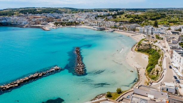 Vista aérea de un pueblo costero con mar azul cristalino, un rompeolas rocoso y edificios a lo largo de la costa.