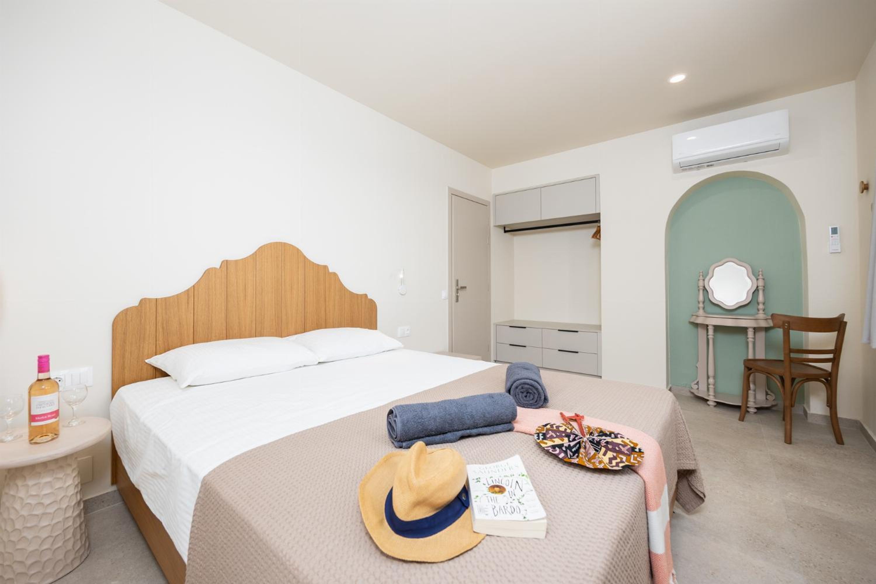 Habitación de hotel con cama de madera, mesita de noche con una botella, vasos, un sombrero y un libro. Hay una silla, un escritorio con un espejo y un equipo de aire acondicionado en la pared.
