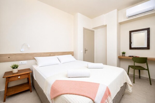 A hotel room with a bed, a small bedside table with a plant, a desk with a chair, and an air conditioning unit.