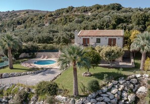 A stone house with a swimming pool surrounded by a stone wall, palm trees, and a hilly landscape.