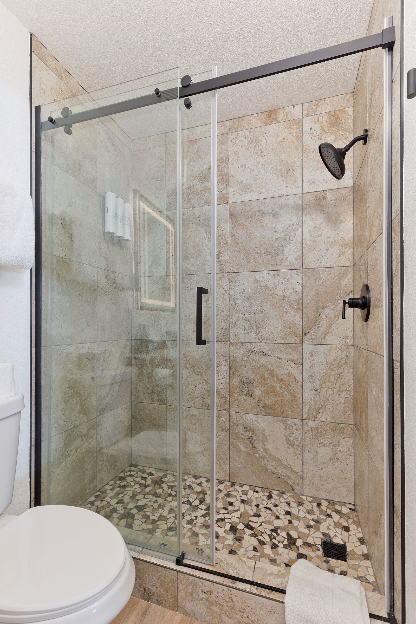 A glass-enclosed shower with a showerhead and a control panel, a tiled floor, and a white toilet.
