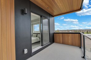 Ein moderner Balkon mit Holzboden, großen Schiebeglastüren, die zu einem Schlafzimmer führen, und Blick auf die Stadtlandschaft.