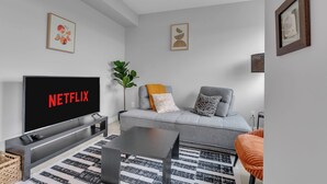 En moderne stue med en grå sofa, et sort sofabord, et fjernsyn der viser 'NETFLIX' og en tæppe med et mønster.