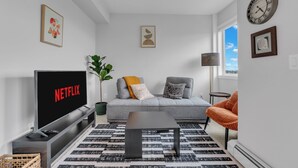 En moderne stue med en grå sofa, et sort sofabord, et fjernsyn der viser 'NETFLIX', og et stort vindue med udsigt.