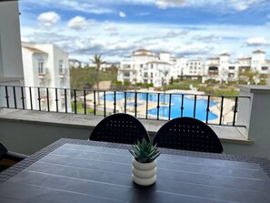 Un balcon donnant sur une piscine et des immeubles, une table avec un pot de fleurs et deux chaises.