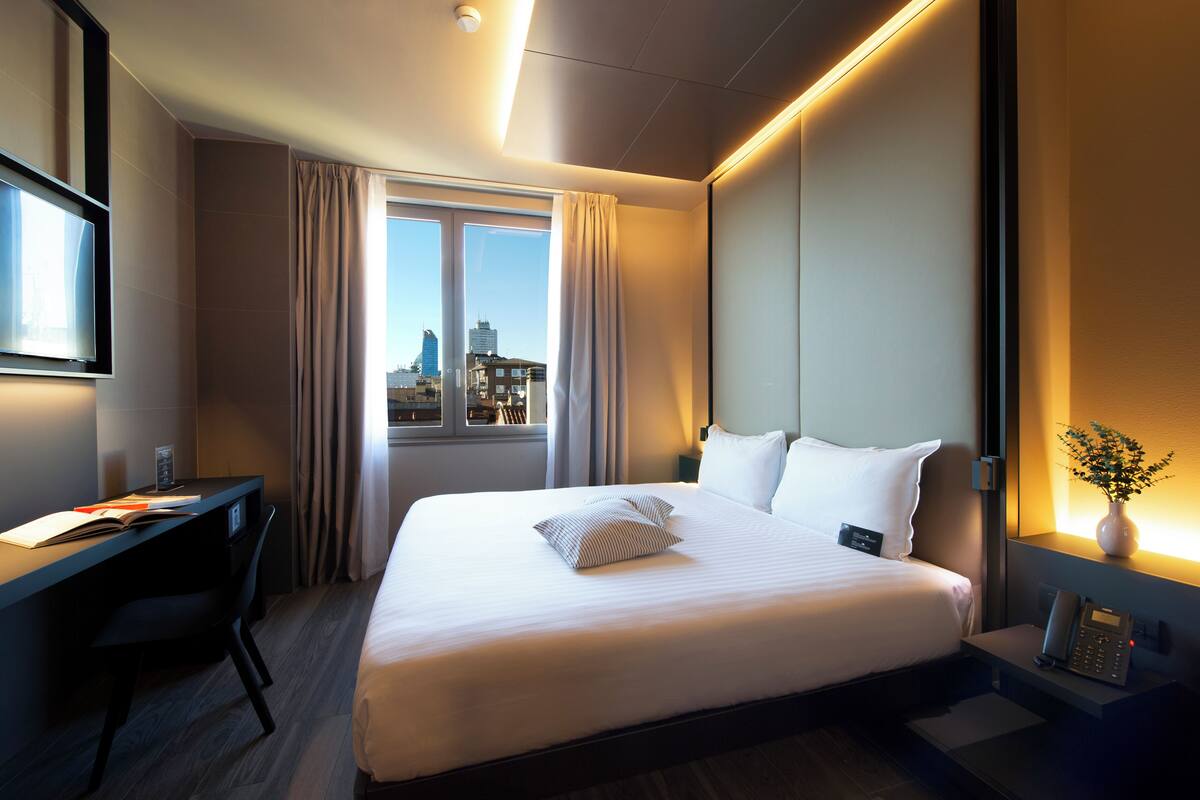 A hotel room with a large bed, a desk, a chair, a window with curtains, and a city view.