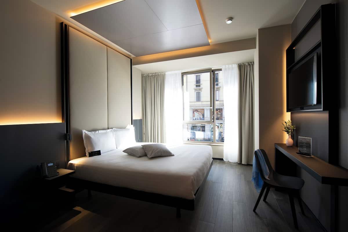 A modern hotel room with a large bed, a desk, a chair, a flat-screen TV, and a window with curtains.