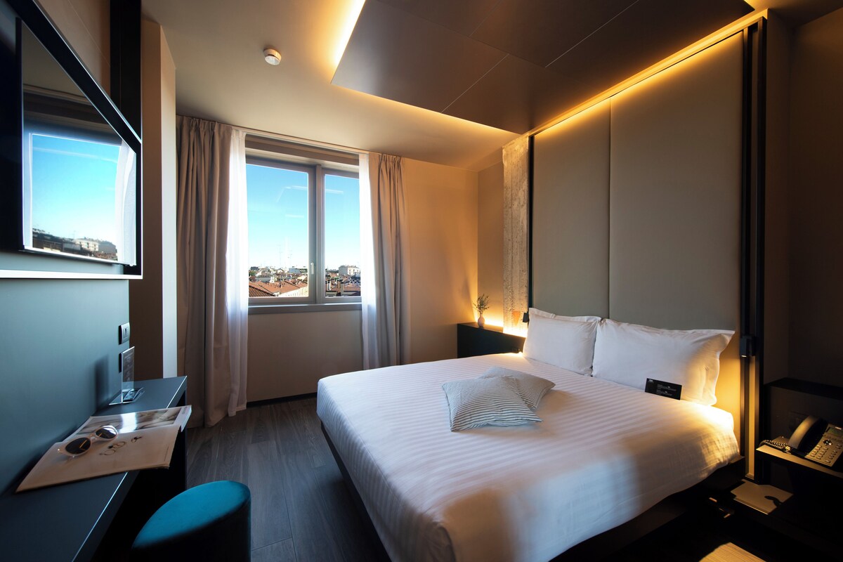 A hotel room with a large bed, a desk with a chair, a nightstand with a lamp, and a window with curtains.