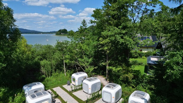 A row of modern, white, cube-shaped mobile homes with large windows, arranged in a grassy area with a lake and trees in the background. - Directly on Lake Wallersee - Cubes - SELF CHECK-IN (Henndorf am Wallersee)