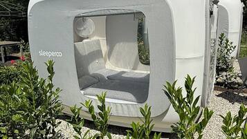 Un remolque habitable blanco con ventana y puerta, rodeado de vegetación.