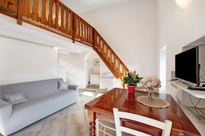 Un salon avec un escalier en bois, un canapé gris, une table à manger en bois avec des chaises et un téléviseur posé sur un meuble.