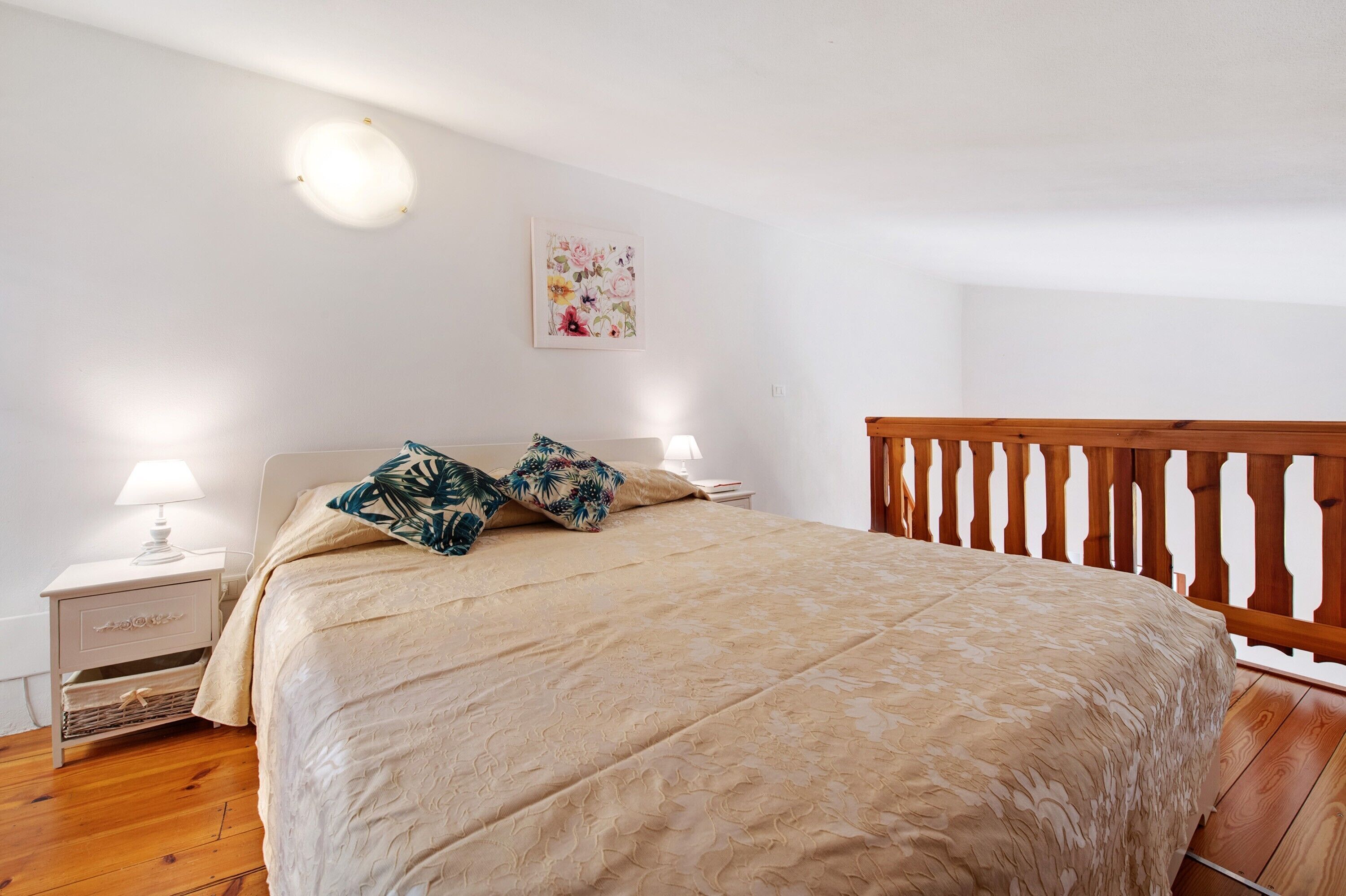A bedroom with a bed, nightstands, a lamp, a wooden railing, and a floral wall decoration.