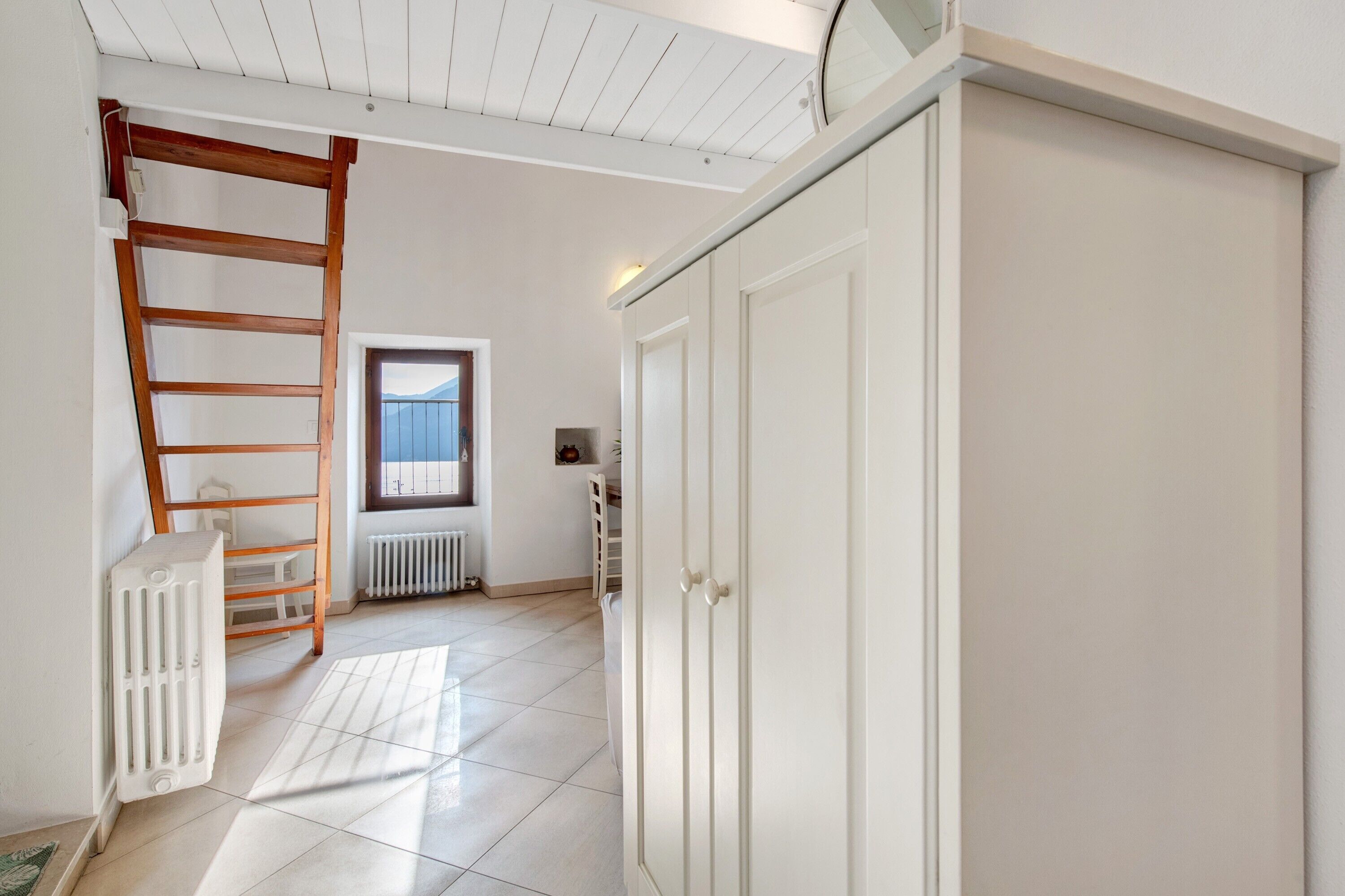 Une pièce avec un radiateur blanc, une échelle en bois, une armoire blanche et une fenêtre donnant sur un beau paysage.
