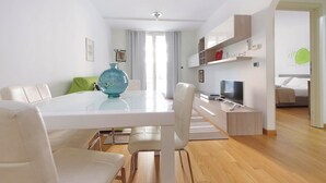 A modern living room with a glass dining table, white sofas, a TV, and a kitchen area with wooden cabinets. - 4BNB - Porta Portese Cozy Apartment (Rome)