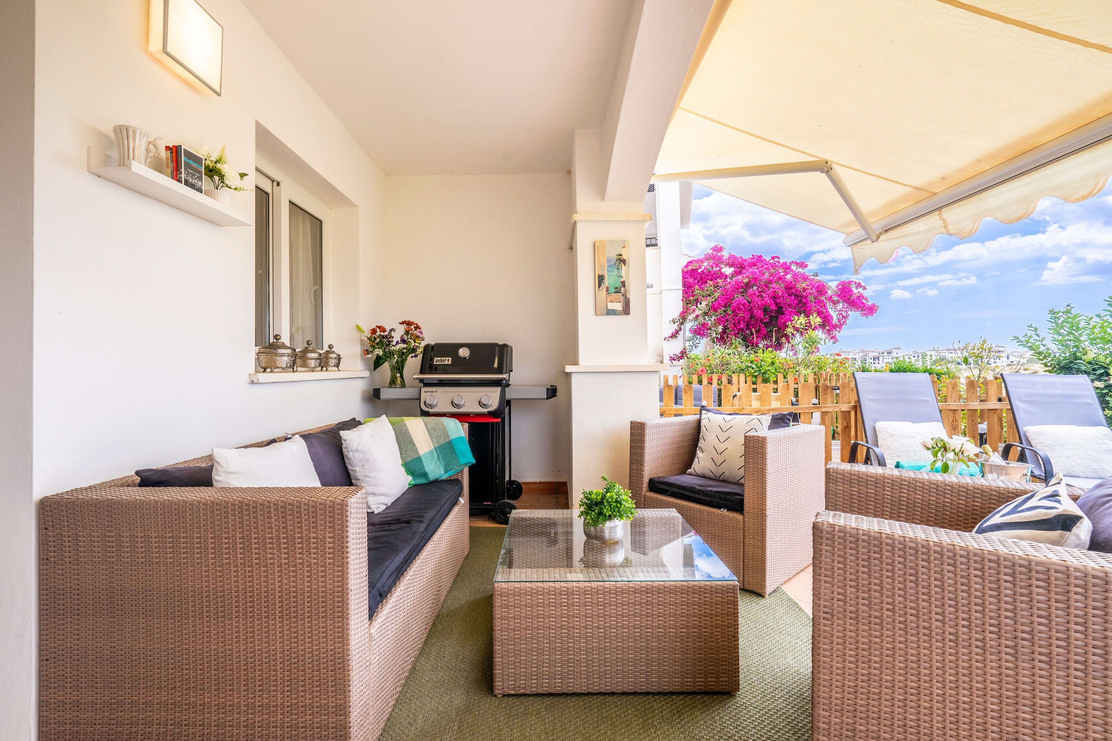 Een gemeubileerd terras met rieten meubilair, een salontafel met glazen blad en een barbecue.
