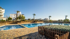 Zona de piscina con suelo de piedra, palmeras y edificios al fondo.