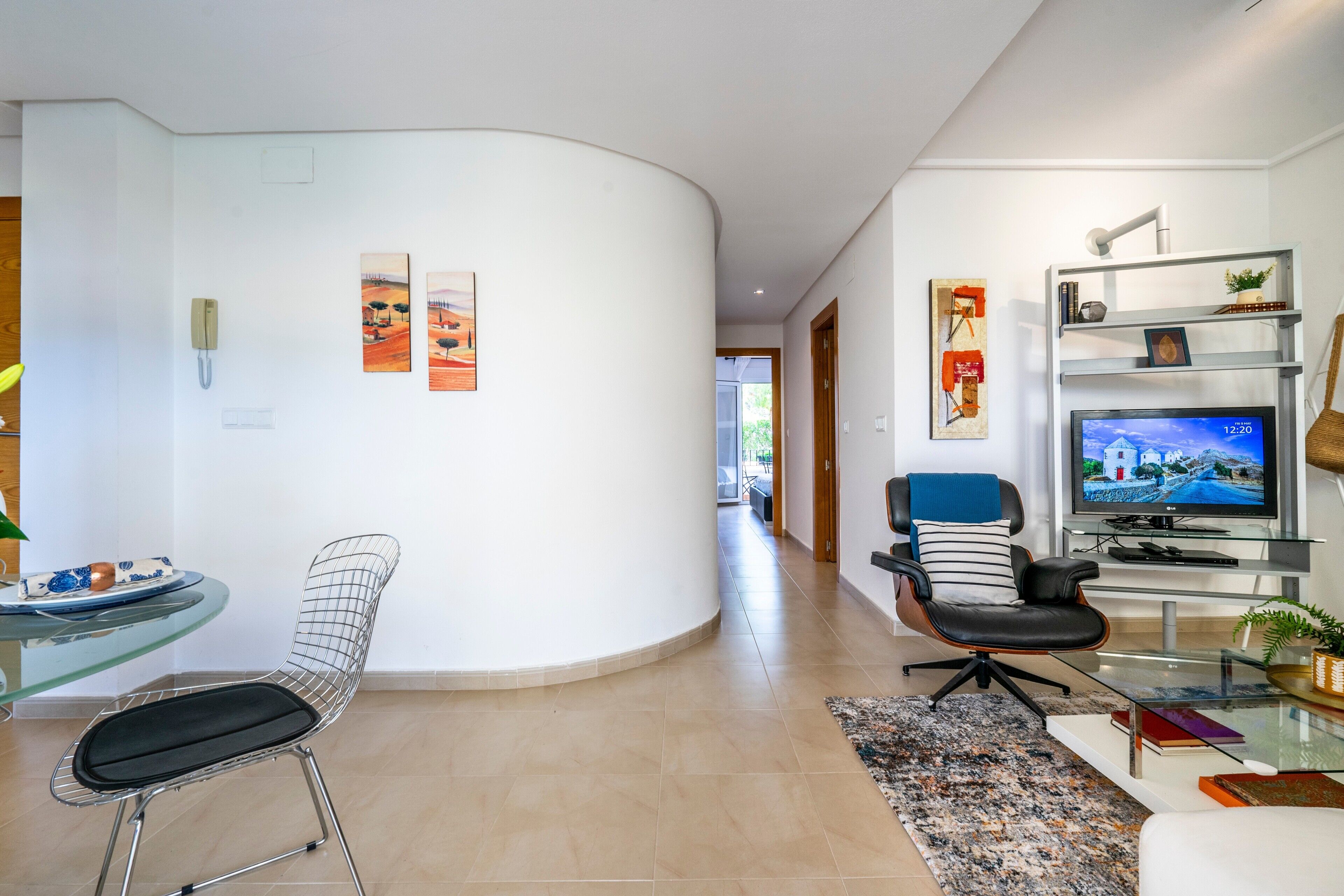 A modern living room with a curved white wall, a glass table with a chair, a black leather sofa, a television, and a bookshelf.