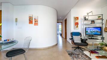 A modern living room with a curved white wall, a glass table with a chair, a black leather sofa, a television, and a bookshelf.