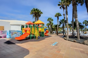 A playground with slides and climbing structures, palm trees, and a tiled walkway.