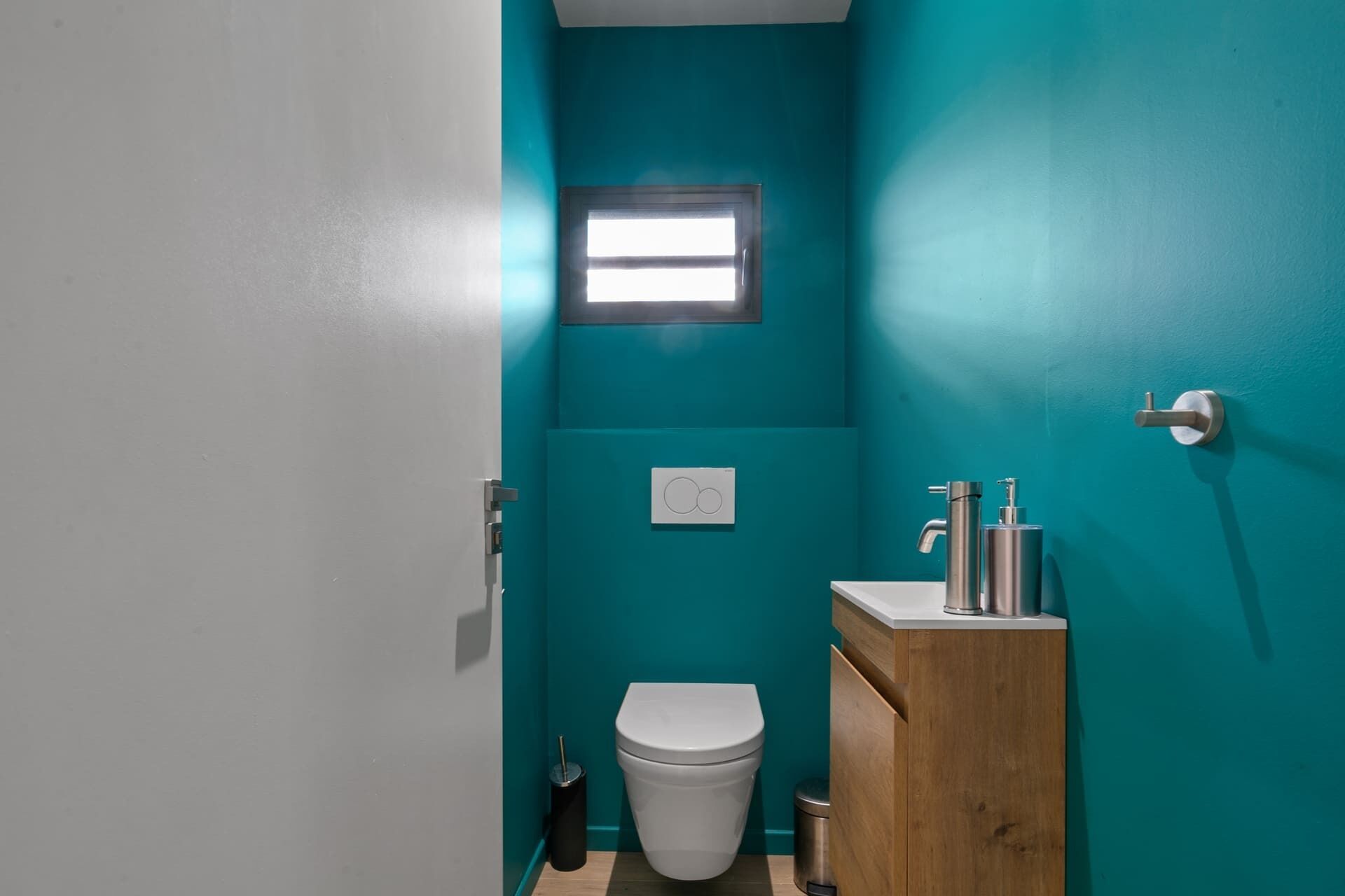 Une petite salle de bain aux murs de couleur turquoise, avec des toilettes blanches, un meuble-lavabo en bois et une fenêtre.