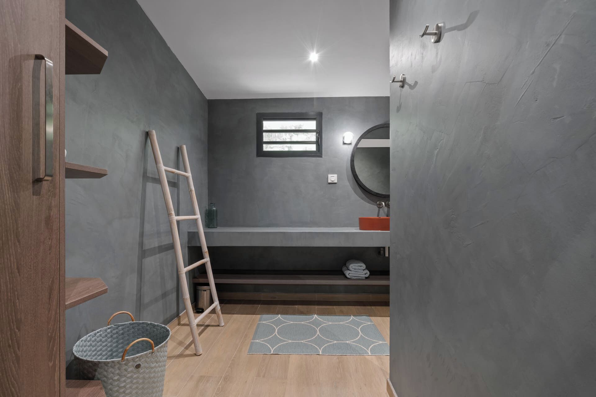 A bathroom with a wooden door, a ladder, a small window, a round mirror, and a grey patterned rug.