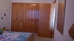 A bedroom with a wooden wardrobe, a bed with a floral bedspread, and a small table with a vase and a plant. - S+1 at Résidence Ghassen (Ariana)
