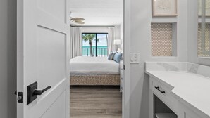 A hotel room with a bed, a view of the ocean, a ceiling fan, and a bathroom with a white countertop.