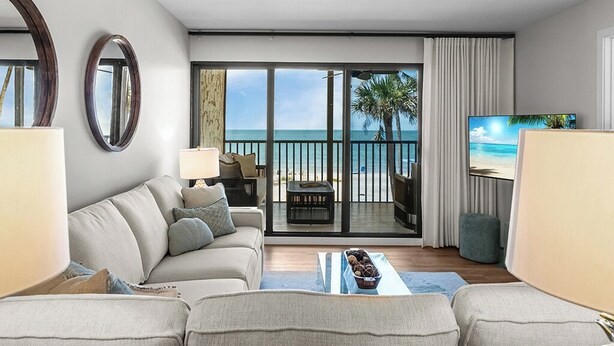 A modern living room with a sofa, a coffee table, and a TV. The room has large windows with a view of the ocean and palm trees.
