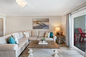 Een woonkamer met een hoekbank, een salontafel, een schilderij, een lamp en uitzicht door een glazen deur.