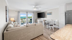 A living room with a sofa, a dining table with chairs, a ceiling fan, a TV, and a view of the ocean.