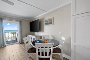 A dining area with a round table, white chairs, a flat-screen TV mounted on the wall, and a view of the beach through a sliding door. - 1bd 1.5ba 3rd Floor Summit Beach Front Sleeps 5 (Panama City Beach)