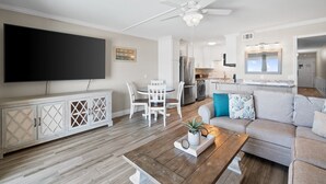 A modern living room with a flat-screen TV, a dining area, a kitchen, and a ceiling fan.