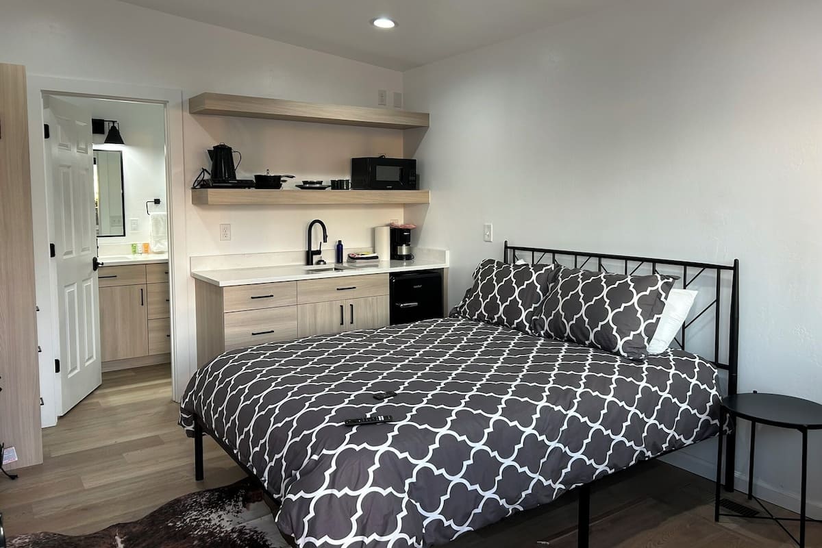 A bedroom with a bed, a bedside table, a small desk, a built-in shelf with kitchen items, and a door leading to a bathroom.