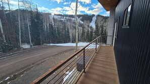 A wooden deck with a metal railing overlooking a snow-covered road and forested mountains.